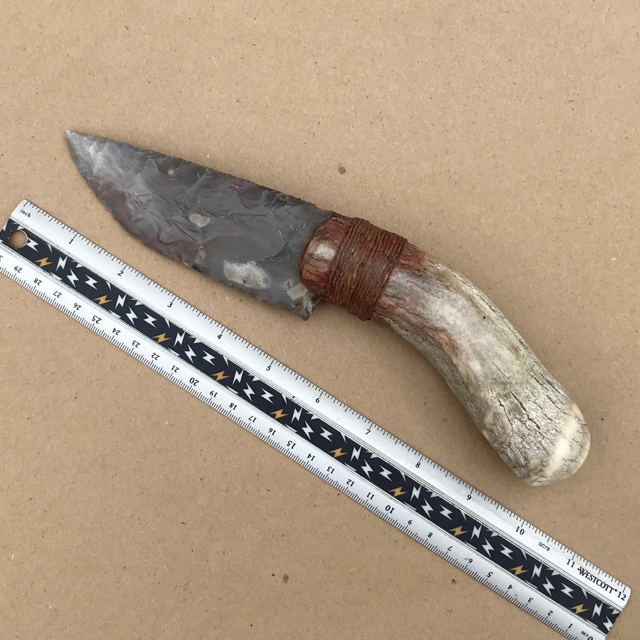 Flint bladed knife with antler handle next to a ruler on a brown background