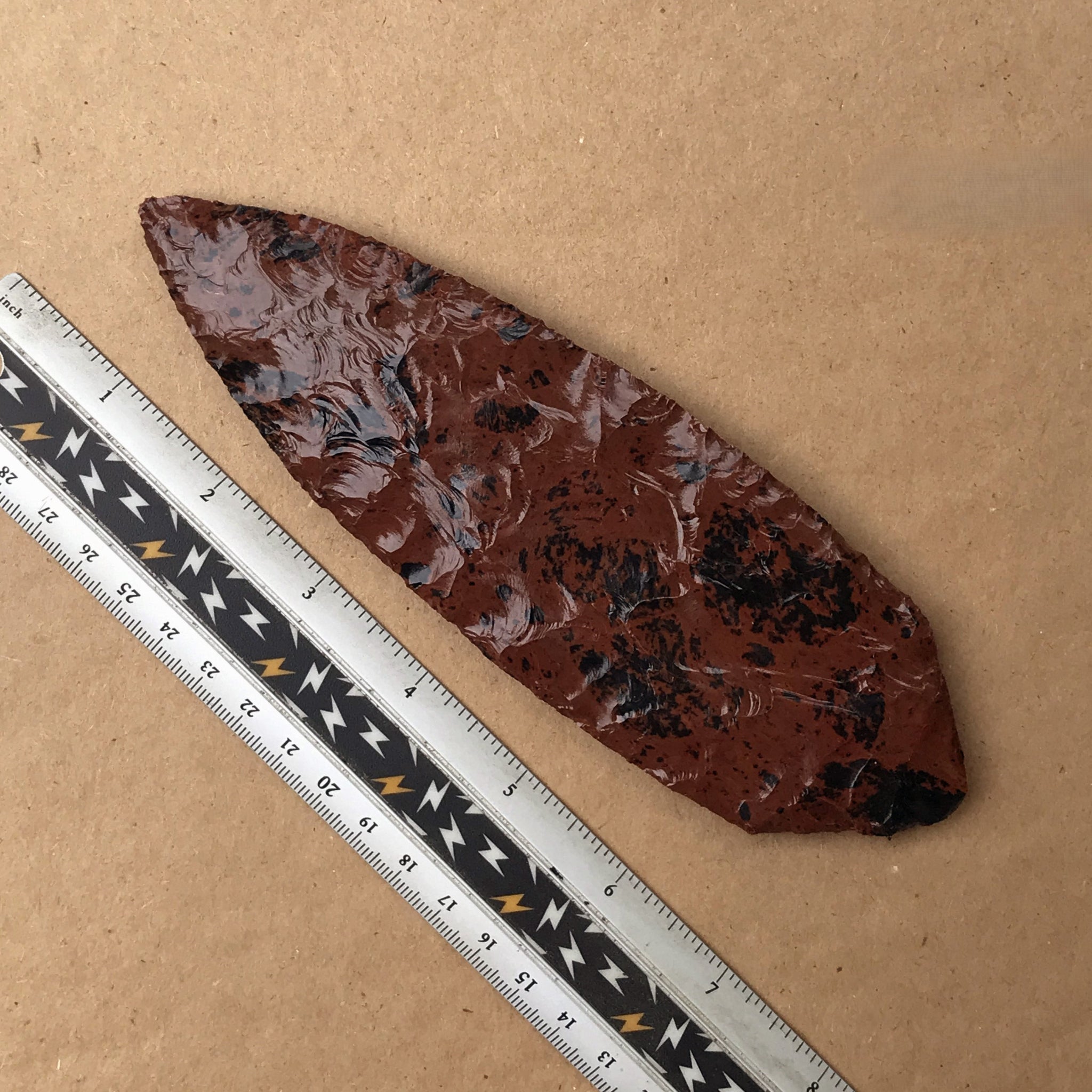 Mahogany obsidian blade with a ruler for scale on a brown background