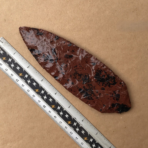 Mahogany obsidian blade with a ruler for scale on a brown background