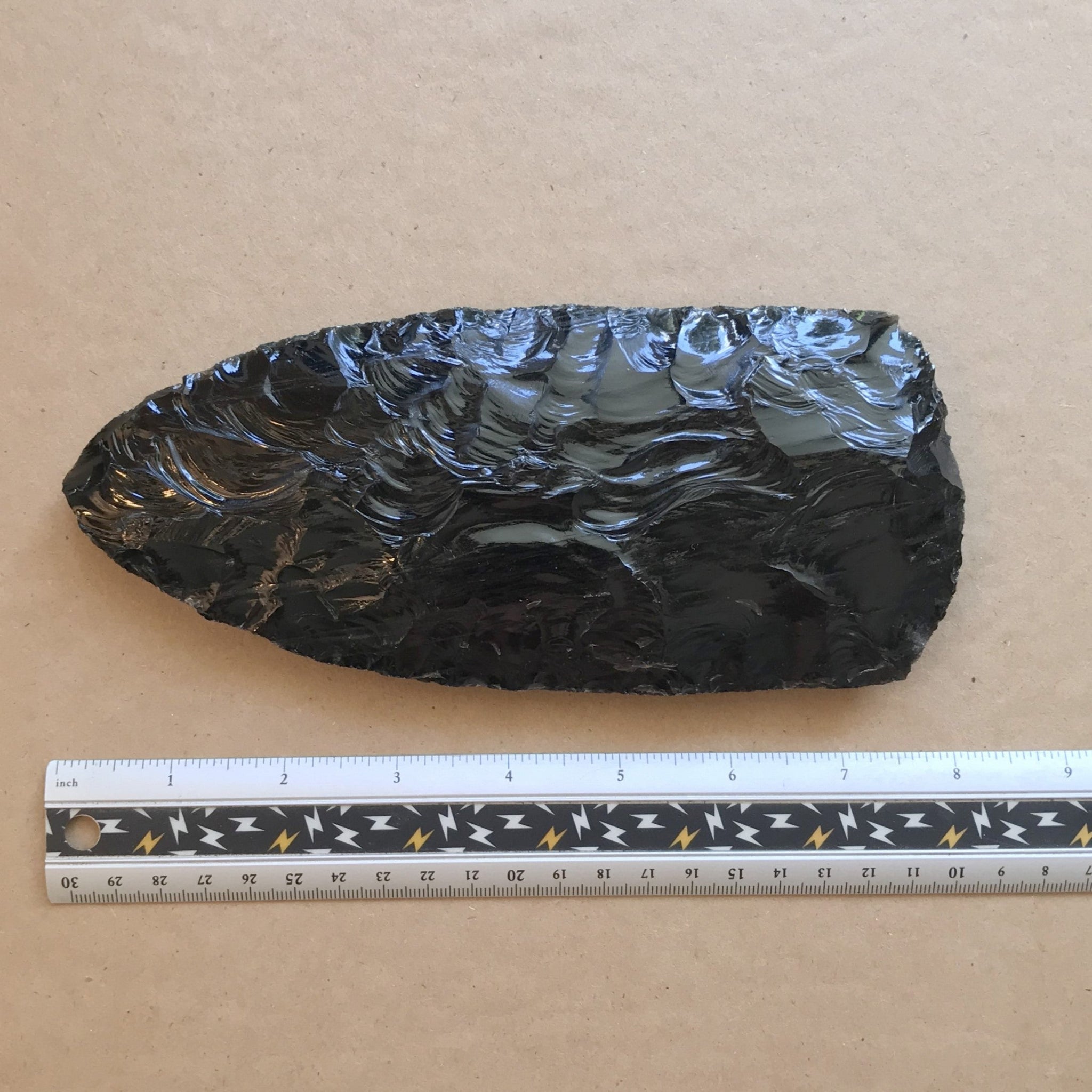 Oversized black obsidian stone axe head with a ruler for scale on a brown kraft paper background