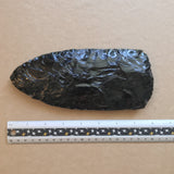 Oversized black obsidian stone axe head with a ruler for scale on a brown kraft paper background