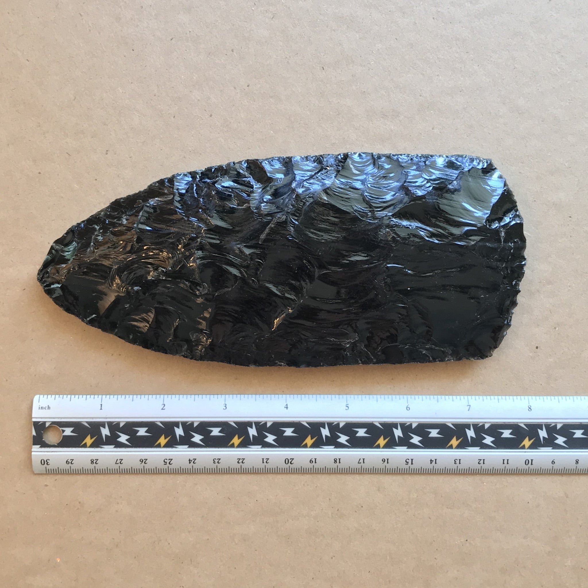 Black obsidian stone axe head with a ruler for scale on a brown kraft paper background