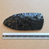 Black obsidian stone axe head with a ruler for scale on a brown kraft paper background