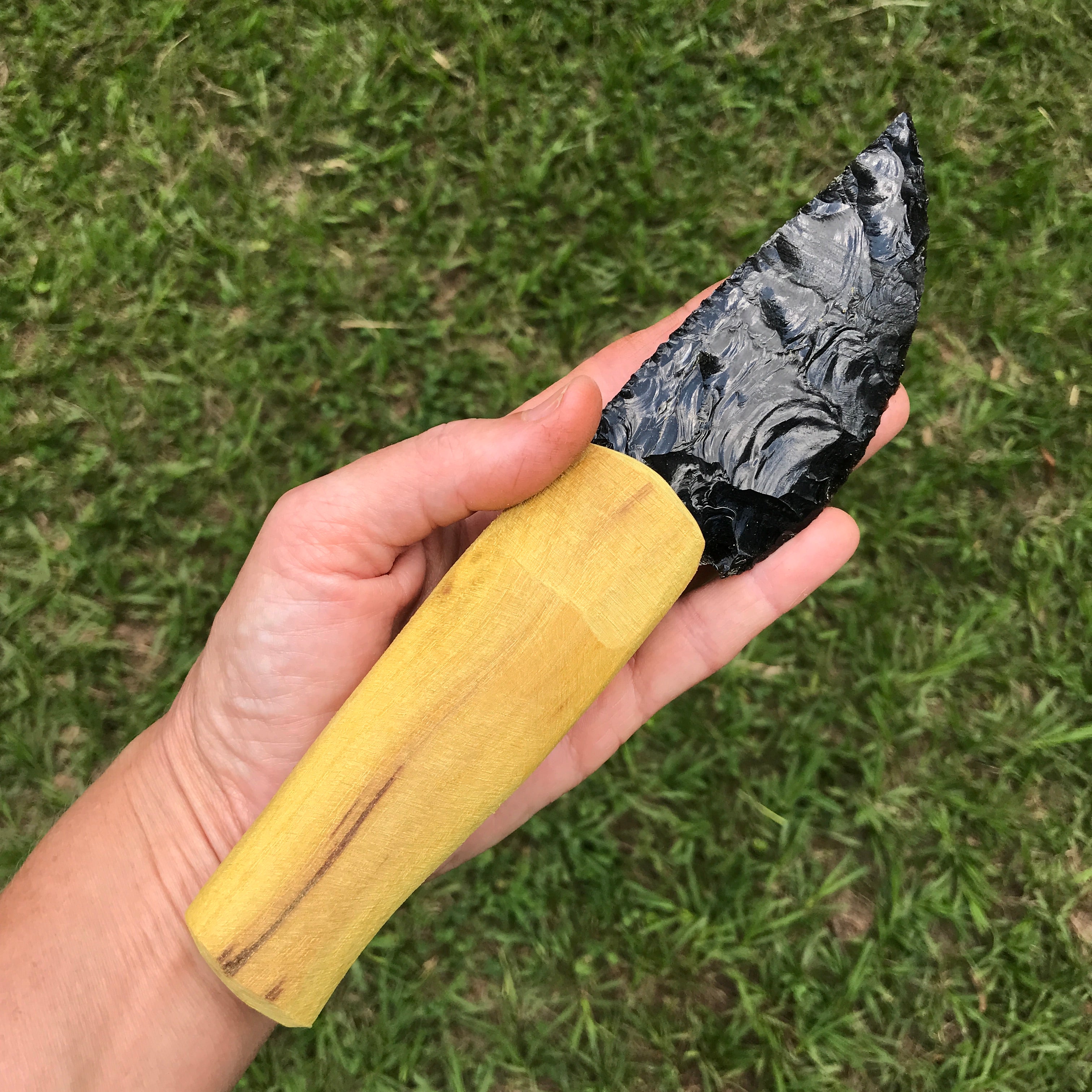 Hand holding a black obsidian blade with an Osage orange handle kit components against a grassy background