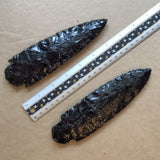 Reverse of two black obsidian blades with a ruler for scale on a brown background